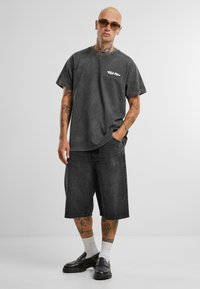 Sötétszürke, oversized T-shirt logóval, térdig érő sötét farmernadrággal párosítva. Fekete loaferek és fehér zoknik teszik teljessé az öltözéket.