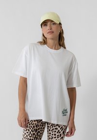 Valge puuvillane T-särk, millel on mugav lõige ja roheline "Hea Karma Klubi" grafika. Kombineeritud leopardimustriliste retuusidega ja heleda kollase mütsiga.