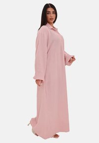 Il modello si trova di lato indossando un lungo abito rosa con bottoni e colletto. Mostra una posa rilassata, mettendo in risalto il tessuto e lo stile.