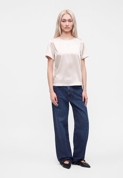 Jeune femme aux longs cheveux blonds portant un haut en satin beige clair à manches courtes, un jean large bleu foncé et des chaussures noires à bouts pointus.