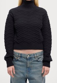 Pull noir en maille avec col montant, présentant une texture ondulée et des poignets côtelés, associé à un jean taille haute délavé clair.