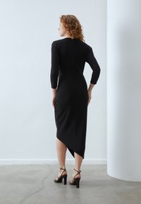 Schwarz formschönes Kleid mit Ellenbogen-langen Ärmeln, asymmetrischem Saum und glatter Textur. Getragen mit schwarzen, strappy High Heels.