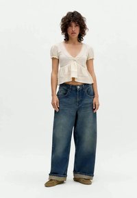 Blusa branca de mangas curtas com bainha peplum e decote em V, combinada com jeans oversized de ganga azul escura. O modelo usa sapatos slip-on castanhos.