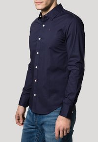 Navy blue long-sleeve shirt with a button-down collar and white buttons. Features a small logo accent on the chest and a smooth texture.