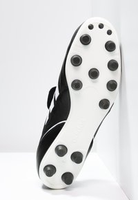 Chaussure de football noire avec une tige texturée, accents blancs et une semelle blanche plate dotée de crampons ronds noirs pour une meilleure adhérence.