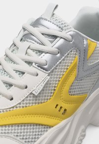 Λευκό και γκρι αθλητικό παπούτσι με κίτρινες λεπτομέρειες, με ύφασμα από δίχτυ, λευκά κορδόνια και χοντρή λευκή σόλα, απεικονισμένο σε κοντινή γωνιακή προβολή.