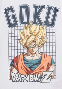 T-shirt blanc avec un graphisme de Goku de Dragon Ball Z, ayant des cheveux blonds en piques, une carrure musclée et une tenue bleu et orange.