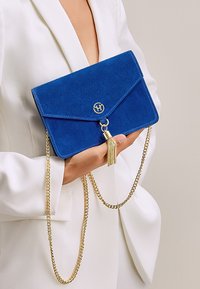 Borsa a pochette blu in suede con tracolla in catena dorata, patta triangolare e una frangia decorativa. Logo dorato al centro sul davanti.