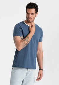 Granatowa koszulka z białym okrągłym dekoltem, krótkimi rękawami i luźnym krojem. Posiada widoczne wzory tatuaży na ramieniu.