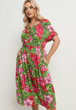 Woman wearing a green, pink, and red floral off-shoulder dress with short sleeves, hands in pockets, and gold bracelets on her right wrist.