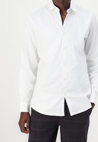 Camicia bianca a maniche lunghe con colletto abbottonato, caratterizzata da una texture liscia e un taglio dritto. I bottoni sono di colore crema.