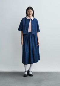 Mujer de pie con falda azul bordada y chaqueta corta sobre una camisa rosa con cuello, lleva calcetines blancos y zapatos negros tipo Mary Jane.