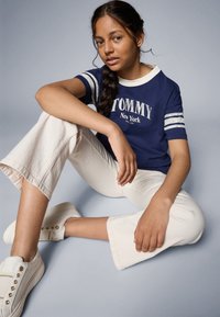 Marineblå T-skjorte med hvit tekst og striper, kombinert med hvite, korte bukser og sneakers. Modellen har langt hår i flette og sitter foran en blå bakgrunn.