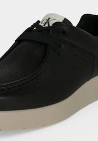 Μαύρο δερμάτινο casual παπούτσι με μπεζ σόλα, μαύρα κορδόνια και λευκή ετικέτα στη γλώσσα με το λογότυπο CK Calvin Klein.