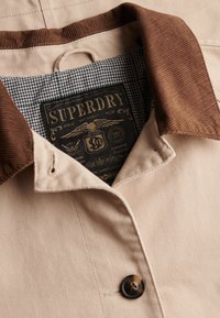 Chaqueta beige con cuello de pana marrón, botón negro, forro interior de cuadros y etiqueta negra de la marca Superdry dentro del cuello.