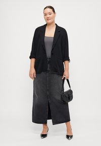 Blazer noir avec les manches retroussées, top gris, et une jupe maxi en denim noire taille haute avec une fente à l'avant. Sac à main et chaussures noires.
