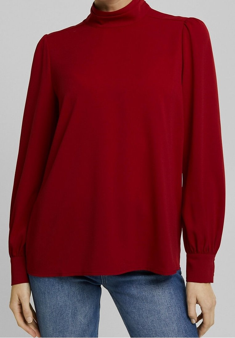 Rød langærmet top med høj hals, plisserede skuldre, glat tekstur og løs pasform. Ærmerne samlet ved mansjetten, parret med jeans.