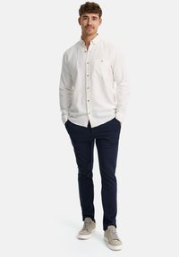 Camisa de botón color beige claro con un bolsillo en el pecho, combinada con pantalones ajustados azul marino y zapatillas gris claro. Tejido suave, diseño clásico.