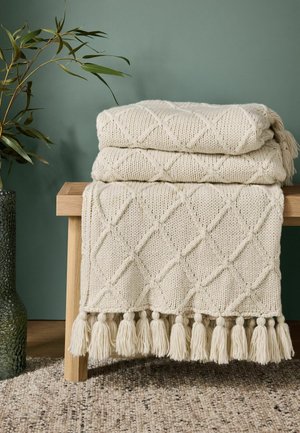 Coperta lavorata a maglia color crema con motivo a diamante e nappine piegata su una panchina in legno chiaro accanto a una pianta con foglie verdi in un vaso.