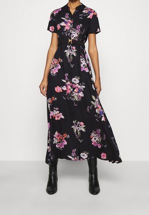 Robe maxi noire à manches courtes avec imprimé floral, col et ceinture, portée avec des bottines noires, debout devant un fond clair uni.