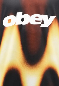 Έντονο λευκό κείμενο "obey" σε ύφασμα με σκοτεινό φόντο και διαβαθμισμένες φλόγες σε κίτρινο, πορτοκαλί και μαύρο χρώμα.