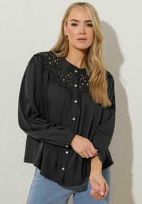 Svart långärmad blus med rund, spetsprydd krage, som har utskärningar och framknäppning. Mjuk, flödande tyg.