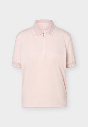 Persona con camiseta polo de manga corta color rosa claro, cinturón y pantalones marrones, calcetines blancos, zapatillas blancas, de pie frente a un fondo claro y liso.