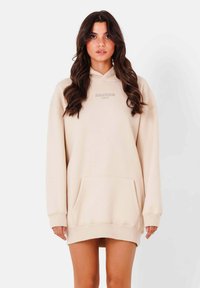 Vestido sudadera beige de gran tamaño, confeccionado en una tela suave, que cuenta con un bolsillo frontal, capucha con cordón y un sutil texto bordado en el pecho.