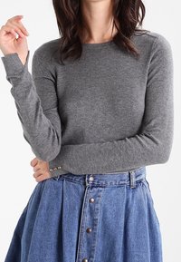 Pull gris à manches longues en tissu tricoté, doté d'un col rond et de détails subtils de couture, associé à une jupe bleue à boutons sur le devant.