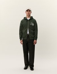 Zelená mikina na zips s predným vreckom a bielym logom na ľavej hrudi, skombinovaná s tmavými džínami a čiernymi topánkami, doplnená béžovou čiapkou.