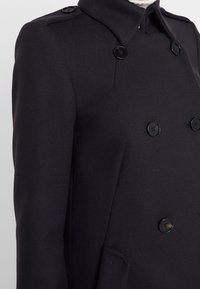 Abrigo de lana azul marino con diseño de doble botonadura, botones negros y cuello alto. Tejido texturizado con bolsillos laterales y hombreras.
