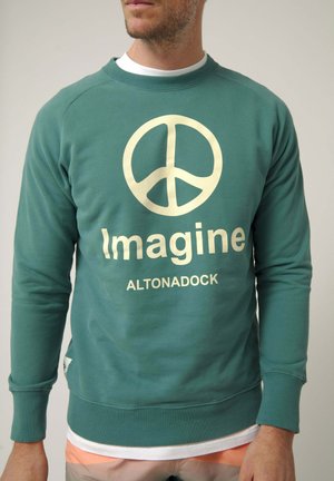 Hombre que lleva una sudadera verde azulado con un gran símbolo de la paz blanco y las palabras "Imagine ALTONADOCK" en el frente.