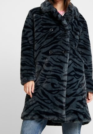 Kvinde iført en mørkegrå og sort tigerstribet imiteret pelsjakke med dobbeltradede knapper over blå jeans og en lyserød top.