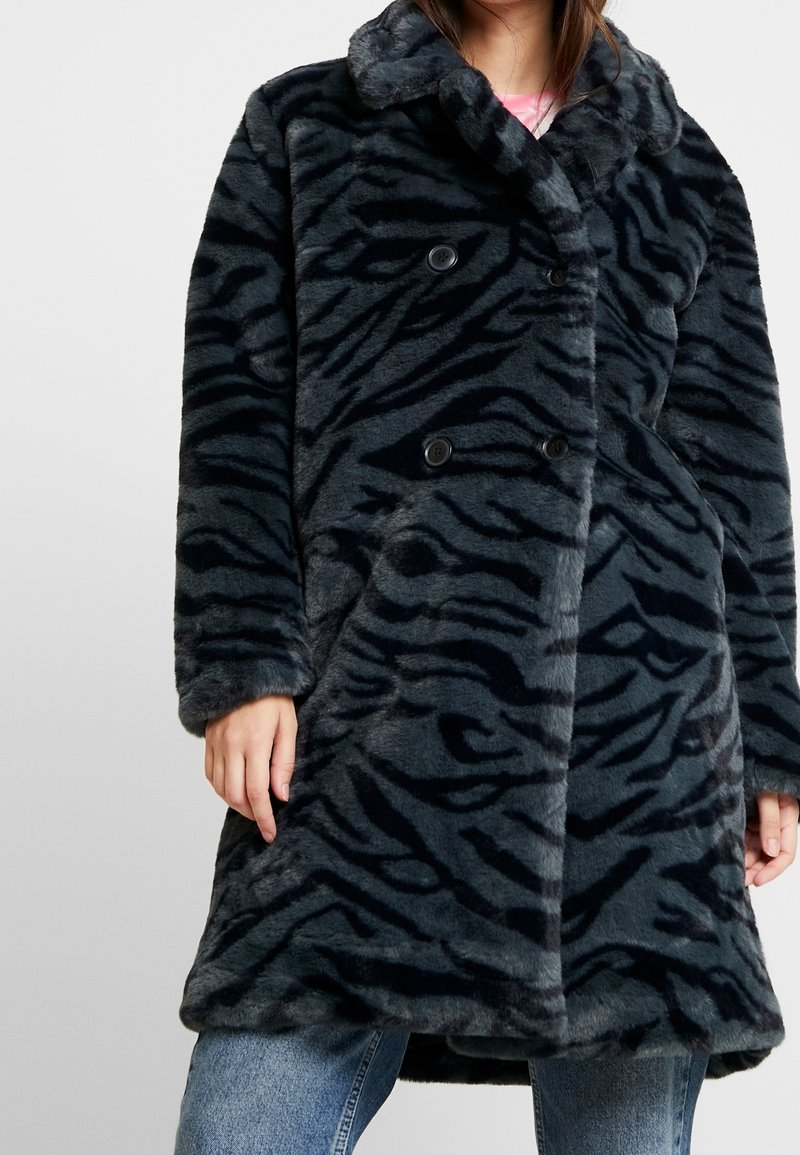 Femme portant un manteau en fausse fourrure à rayures de tigre gris foncé et noir avec des boutons croisés, sur un jean bleu et un haut rose.