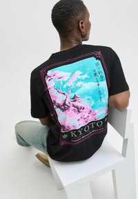 Sort t-shirt med et farverigt kunstværk af kirsebærblomster og en pagode, med "KYOTO" trykt i pink nederst. 100% bomuldsstof.