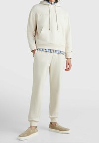 Sudadera beige con logo en relieve, bolsillo tipo canguro, combinada con pantalones de chándal beige a juego. Camisa de rayas azul claro por debajo y zapatos de slip-on en color beige.