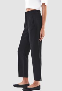 Zwarte tailored broek met een hoge taille, voorzien van voorplooien, zijzakken en een enkellange snit. Soepele stoffen textuur.