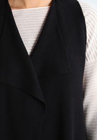Chaleco negro de gran tamaño con cuello chal, superpuesto a una blusa texturizada de un tono beige claro, que exhibe un acabado de punto suave.