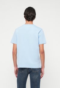 Άνδρας με κοντά σκούρα μαλλιά, φορώντας απλό ανοιχτό μπλε πουκάμισο κοντομάνικο και σκούρα μπλε τζιν, στέκεται στραμμένος με πλάτη σε λευκό φόντο.