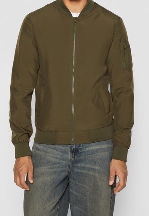 Man wearing olive green bomber jacket with front zipper and zippered pocket on left sleeve, paired with blue jeans.