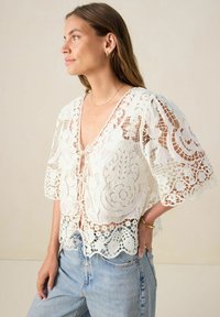 Weiße Spitzenbluse mit V-Ausschnitt, dekorativen Mustern und kurzen, schwingenden Ärmeln. Verfügt über einen Bindeband-Detail vorne und einen gewellten Saum.