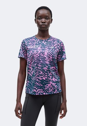 Ženska v roza in mornarsko modri športni majici s kratkimi rokavi z vzorcem ter črnih pajkicah, stoji pred enobarvnim svetlim ozadjem.