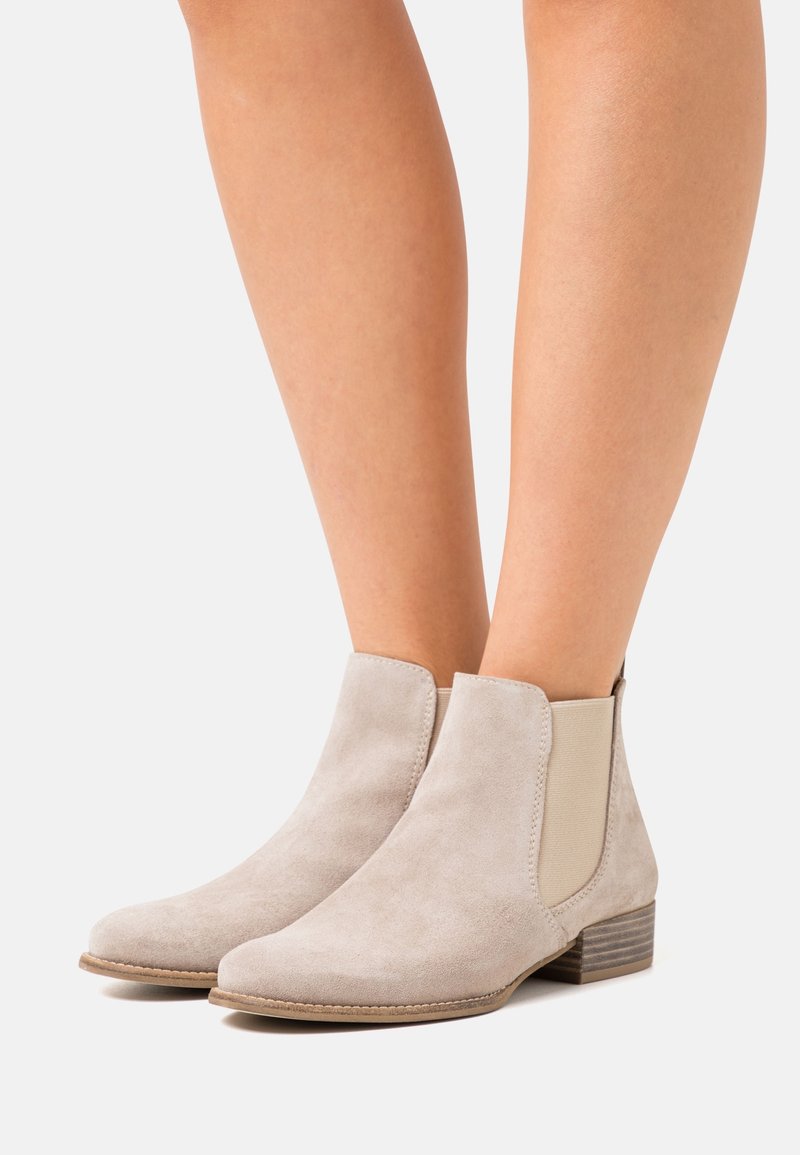 Bottines beige en daim avec panneaux latéraux élastiques et petits talons empilés, portées sur des jambes nues sur fond blanc.