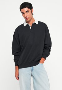 Svart langermet polo-shirt med kontrasterende hvit krage, med en avslappet passform og tre-knappslukking, i kombinasjon med lyseblå jeans.