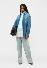 Femme aux longues tresses portant une chemise en jean oversize avec un détail en forme de cœur, un pull à motifs nuages, un jean large clair, tenant un sac noir, debout.
