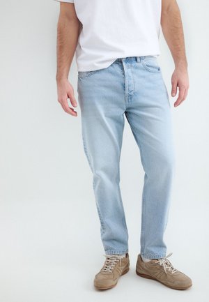 Man draagt lichtblauwe spijkerbroek, wit T-shirt en beige sneakers, staand voor een effen lichte achtergrond.