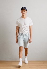 T-shirt blanc, shorts en denim bleu clair et baskets blanches. La personne porte une casquette noire. Texture lisse avec une coupe décontractée. Fond clair.
