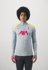 Vaaleanharmaa pitkähihaisen Nike-treenitoppi, jossa on neljännesvetoketju, keltaiset olkapääkoristeet, punainen Liverpool-logo ja suuri punainen AXA-teksti.