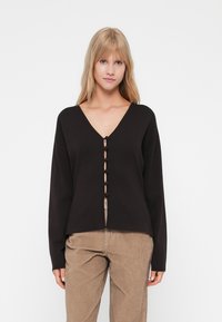 Zwarte lange mouwen cardigan met een V-hals en knoopsluiting aan de voorkant. Glad textuur met een ontspannen pasvorm, gecombineerd met lichtbruine corduroy broek.