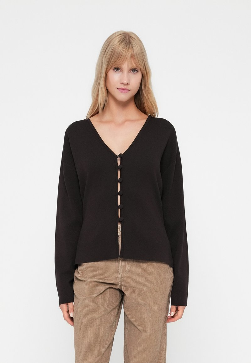 Zwarte lange mouwen cardigan met een V-hals en knoopsluiting aan de voorkant. Glad textuur met een ontspannen pasvorm, gecombineerd met lichtbruine corduroy broek.